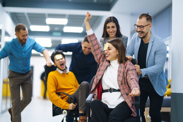 Obraz premium A group of young colleagues are having fun in the office corridor. Young business people ride on office chairs and laugh. Concept of teamwork, relaxation or fun.