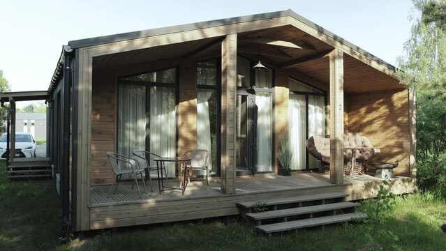 Cozy terrace in the wooden tiny house with warm sunlight on it. Terrace in a small country house in the forest at summer. Table and brown sofa, shadows from trees on the wooden wall, slow motion.