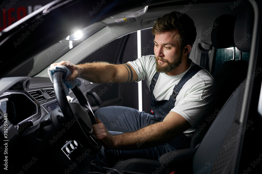 Wall mural Man cleaning interior in car, steering wheel with microfiber cloth. Repair man wipe down the dust inside of auto. Car wash concept. Side View on Concentrated confident male in uniform