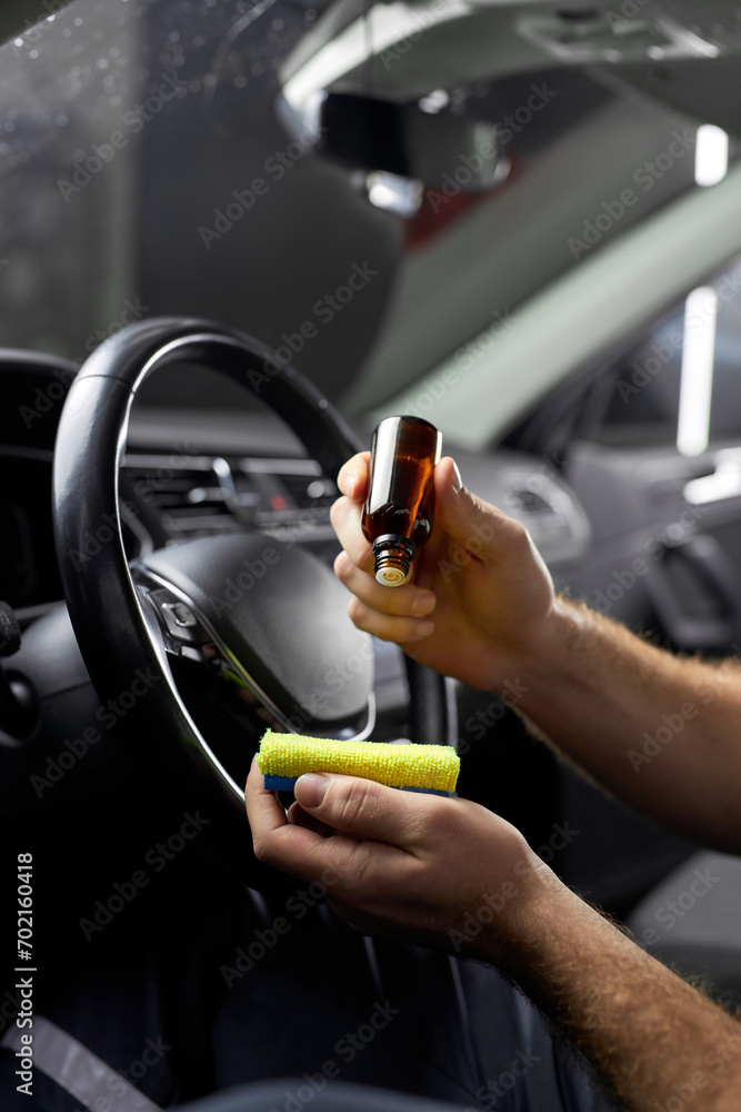 Wall mural professional car wash service worker cleaning car interior with special chemical anti dust liquid. side view on male holding bottle with liquid in hands applying it on rag. close-up hands