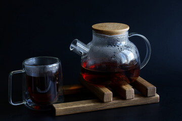 Heat-resistant glass teapot with hot black tea and a heat-resistant mug on a wooden stand for hot...