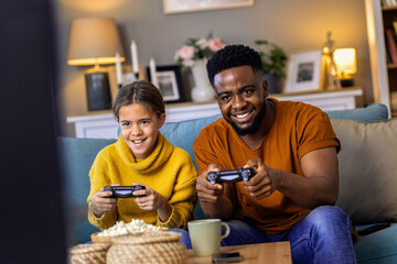 Father and daughter playing video game at home white sitting on sofa in living room.