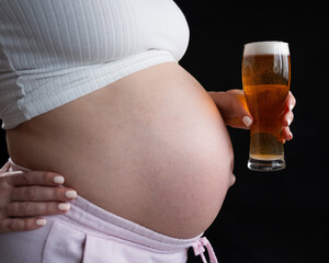 Close-up of the belly of a pregnant woman holding a glass of beer on a black background.