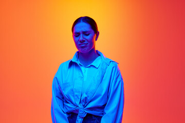 Gloomy dissatisfied brunette girl, student in casual outfit against gradient yellow background in blue neon light. Concept of beauty, youth, human emotions, self-expression, retro.