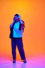 Full-length portrait of overjoyed girl, student dancing while listening music in headphones against gradient studio background in neon light, filter. Concept of beauty, human emotions, self-expression