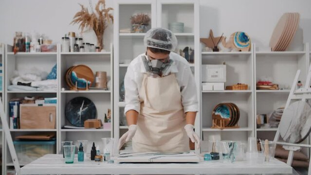 Female artist pours colored art epoxy resin on canvas mixing colors in art studio workshop, creative woman in protective face mask, trendy modern hobby creating beautiful sea waves acrylic paintings