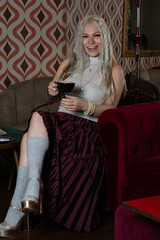 Platinum blonde girl, 25 to 30 years old, wearing a white tank top and a red skirt, gray stockings. With a cup of coffee in her hand, sitting in an antique armchair. Makeup In a 70s style restaurant.