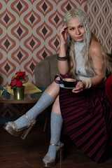 Platinum blonde girl, 25 to 30 years old, wearing a white tank top and a red skirt, gray stockings. With a cup of coffee in her hand, sitting in an antique armchair. Makeup In a 70s style restaurant.