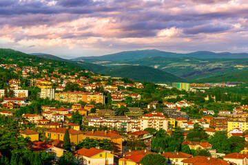 Fototapeta premium panoramic landscape view of green summer city with yellow houses, green forest and mountains above and amazing hills and cloudy sky on background