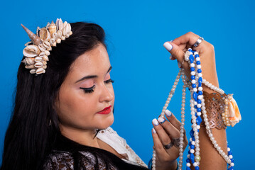 Beautiful, black-haired woman with shell tiara on her head holding and looking at pearl necklaces.
