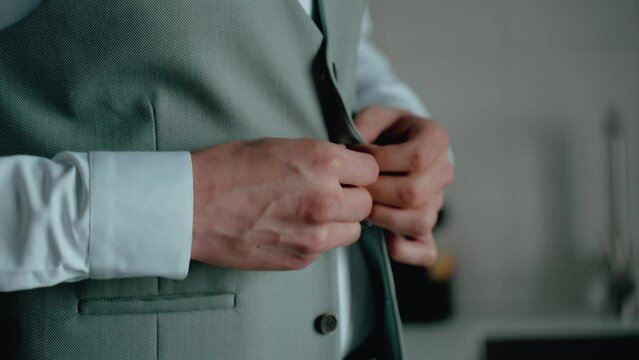 A man in a business suit fastening a button with attention to detail, the focus on his hands and the cuff of the shirt, set against the grey fabric of the suit jacket, conveying a sense of professiona