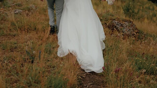 The image captures the lower section of a bride's white flowing wedding dress as she walks through a natural field. The dress's delicate fabric contrasts with the wild, untamed grasses and wildflowers