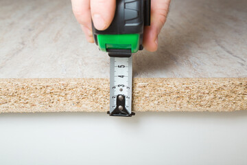 Young adult man hand holding measure tape and measuring edge thickness of new beige tabletop before stove top or sink installation. Closeup. Front view. Kitchen renovation process.