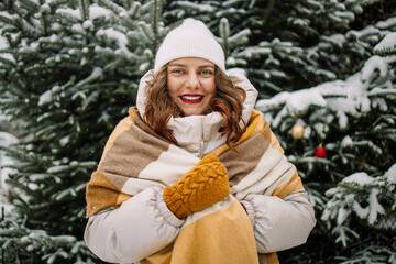 Fototapeta premium Portrait of smiling happy caucasian woman in hat with mittens in winter coat laughing, tossing snow up outside in the city park in winter. High quality photo