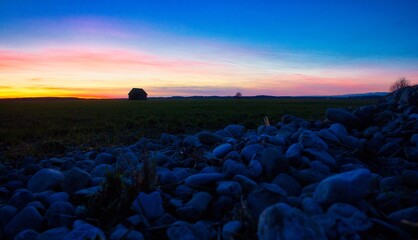 Blaue Stunde - Sonnenuntergang über einer Wiese mit einer Holzhütte im Hintergrund und Steine im...