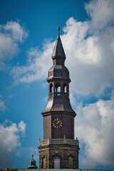 Ein spitz zulaufender, brauner Kirchturm im Sonnenschein, vor blauem Himmel mit weisser Bewölkung