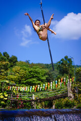 Beautiful Asian girl shows extreme advances by performing pole dance aerial in various positions and spinning stunts on the blue sky around the natural fresh and the waterfall, outdoors.