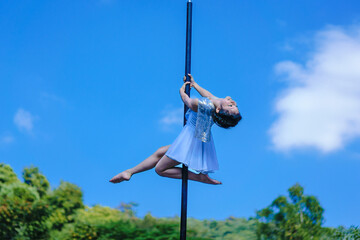 Beautiful Asian girl shows extreme advances by performing pole dance aerial in various positions and spinning stunts on the blue sky around the natural fresh and the waterfall, outdoors. - obrazy, fototapety, plakaty