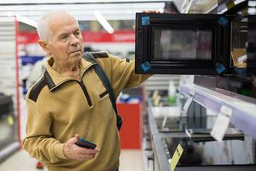 Elderly man looks at a microwave oven on the counter in the showroom of an electrical appliance hypermarket