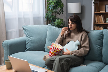 Young woman with stomach ache or menstruation sitting on sofa at home eating popcorn and watching...