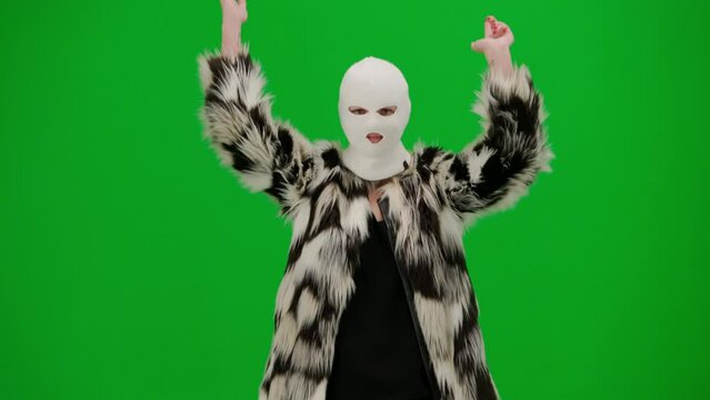 Woman in white balaclava, fur coat and evening dress walking and dancing merrily with her hands up. Woman freak on green background in studio. Fashion trend concept, feminist trend in fashion.