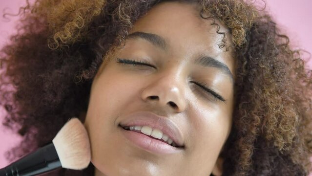 Black woman runs brush over face with eyes closed, smiling enjoying process of applying makeup. African-American happy glad delight girl gets indescribable satisfaction from pleasant touches on skin