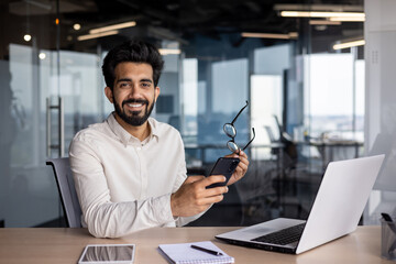 Portrait of a smiling young Indian man sitting in the office at the desk, holding glasses and a mobile phone, looking confidently at the camera