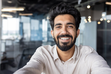 Selfie of a young Indian man standing in the office and smiling at the phone camera. Close-up photo