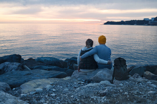 Husband and wife sitting on huge stones near the water and hold hands in the evening.