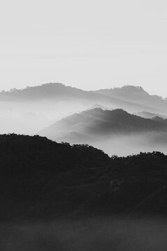 The image is in black and white, emphasizing contrasts between light and shadow. Multiple layers of mountains are visible, each progressively lighter in tone due to atmospheric perspective. Mist or fo
