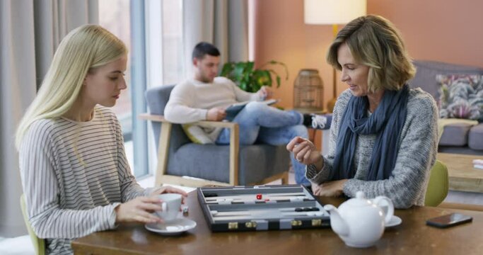 Woman, board game or mom playing backgammon in living room for fun, strategy or bonding at home. Relax, mother and daughter drinking tea in a family together on holiday with dice, challenge or parent