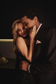 joyful multicultural young couple in elegant evening attires hugging sensually and smiling at home