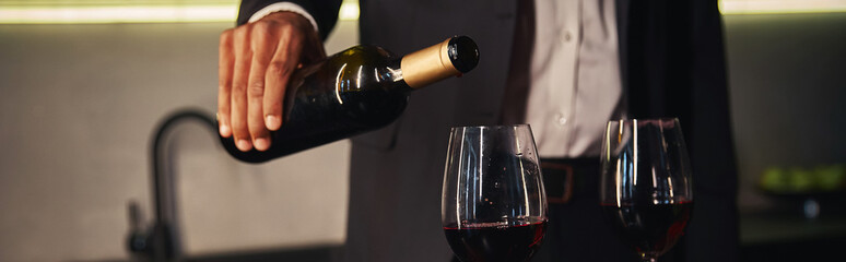 cropped view of young african american man in elegant attire pouring red vine into glasses, banner