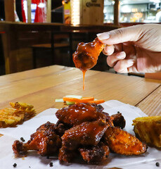 Una mano toma una alita de pollo chorreando salsa BBQ de una pila de alistas sobre una mesa.