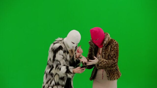 Two women in white and pink balaclavas looking expectantly at a smartphone and getting upset over a loss or bad news. Women freaks in fur coats on the green background of the studio.