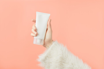 A womans hand in a sweater holds a tube of cream on a peach background. Self care beauty treatment