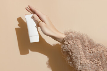 A womans hand in a sweater holds a tube of cream on a beige background. Body care beauty treatment