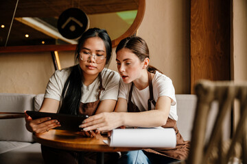 Portrait of two young women cafe staff doing paperwork and using tablet while sitting at table
