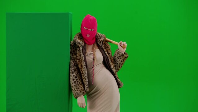 Woman in pink balaclava, tiger coat and dress leaning on green banner and making finger gesture go here. A freak woman with a bat in her hands on a green background in a studio. Fashion trend concept.