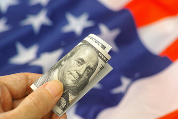 A person holding a one hundred dollar bill above the flag of the USA. Close up.
