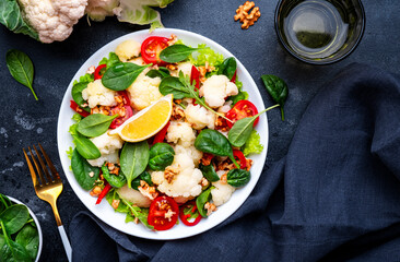 Healthy vegan salad with white cauliflower, baked red paprika, cherry tomatoes and spinach with walnuts, gray table background, top view
