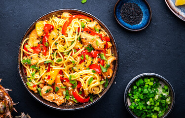 Stir fryed egg noodles with chicken, pineapple, paprika, green onion, soy sauce and sesame seeds in bowl. Asian cuisine dish. Black table background, top view