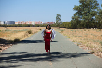 Beautiful young Latin woman in red silk dress and white wings walks along a tarmac road in the countryside outside the big city. The woman makes different poses and expressions.