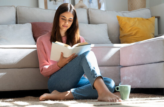 Pretty young woman reading a book and drinking a cup of coffee while sitting on the floor at home.