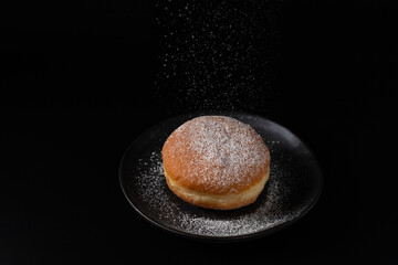 Single fresh baked donut berliner sprinkled with falling sugar powder on retro dark plate on black background. Sweet doughnut closeup.