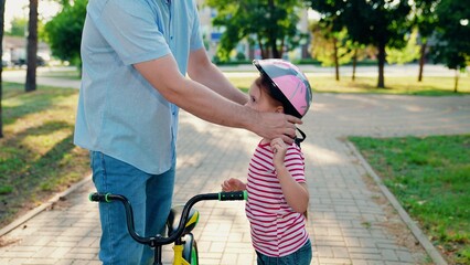 Father teaches kid daughter to ride bike in park. Happy family, little girl in helmet together with...