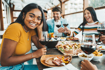 Happy friends drinking red wine sitting at restaurant table - Multiracial young people enjoying...