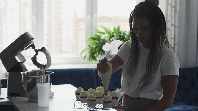 Young woman confectioner puts whipped airy cream from pastry bag to decorate tasty fresh homemade muffin on cooling tray in silhouette opposite bright window