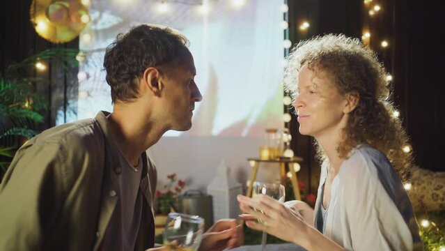 Close-up portrait man and woman with white wine glasses dancing opposite outdoor cinema screen, spending weekend at rest place with pillows and fairy lights in cottage yard