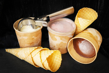 This image shows three cups of ice cream in different flavors: vanilla, strawberry and chocolate. On one of the cups lies a metal ice cream scoop, three empty waffle cones stacked against each other.